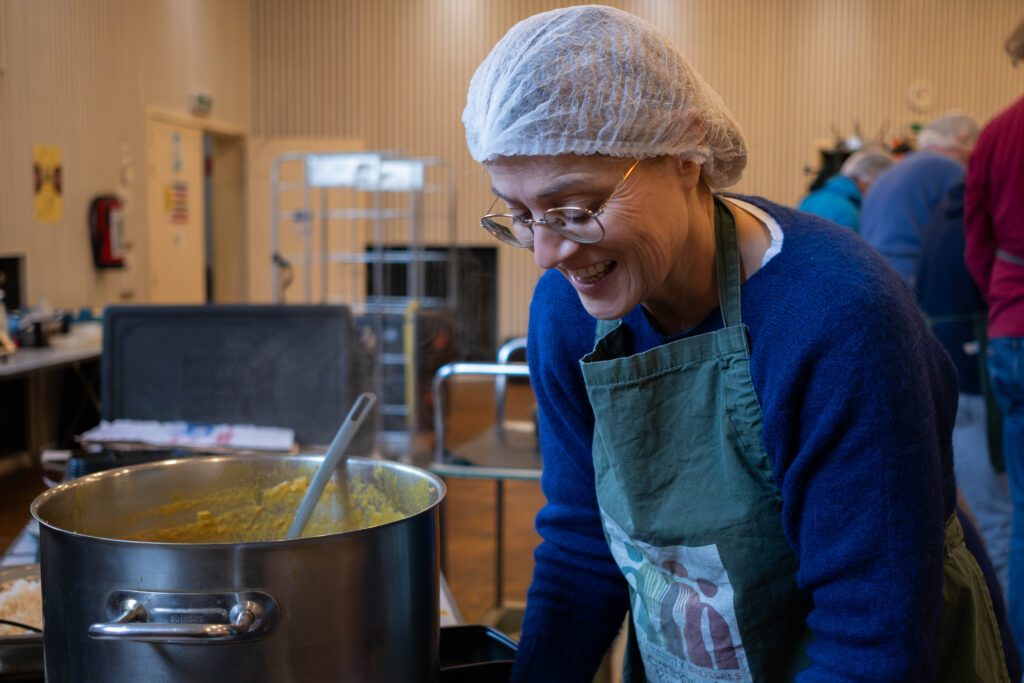 A woman wearing a hairnet and a Community Kitchen apron stands next to a pot of food.